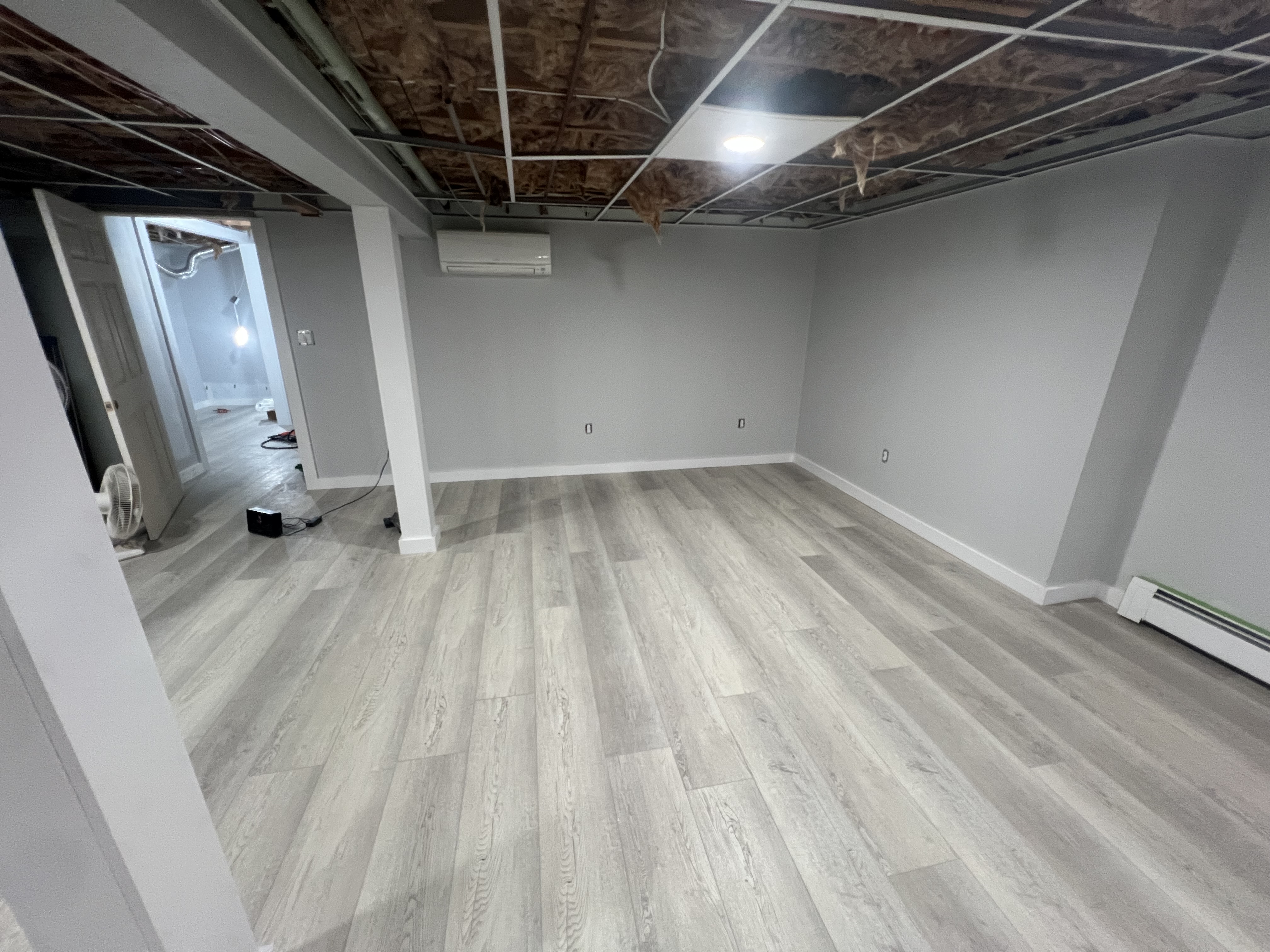 Basement room renovation with finished flooring and open ceiling grid.